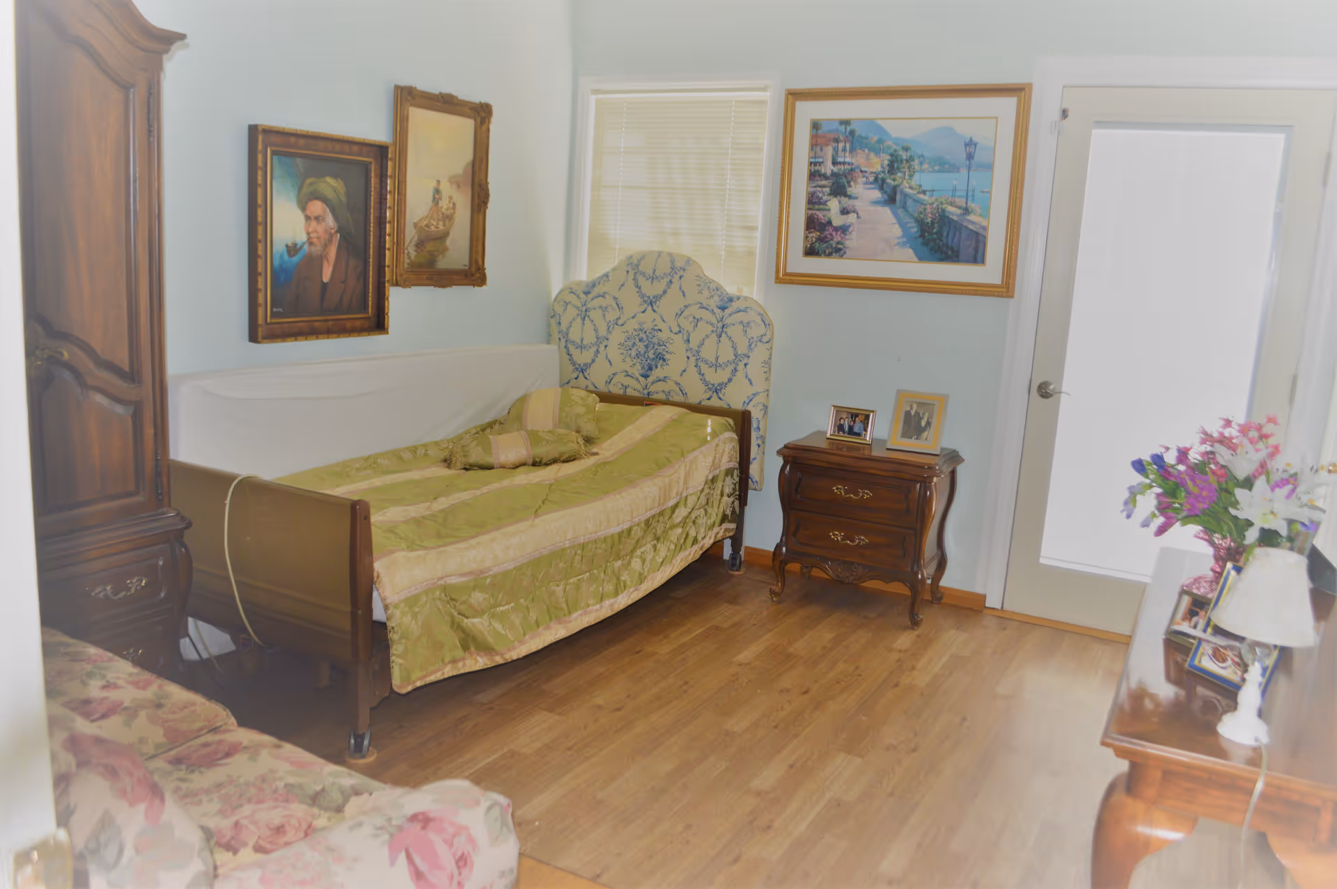 A furnished bedroom with a single bed, wooden nightstand and dresser, framed artwork, and a door with blinds.