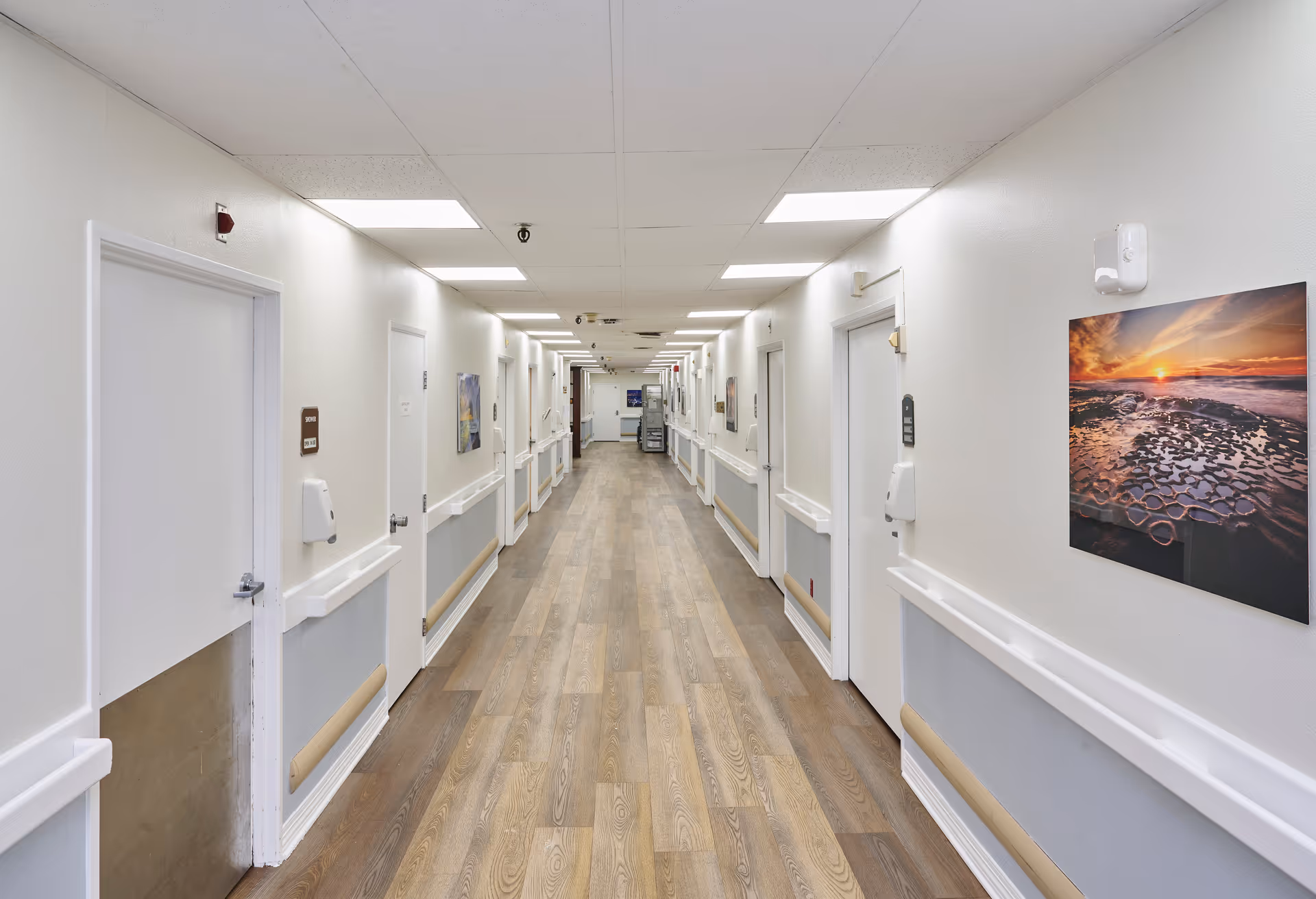 A long, well-lit hallway in a nursing and rehabilitation center with white walls, wooden flooring, handrails on both sides, multiple closed doors, and framed artwork on the walls.