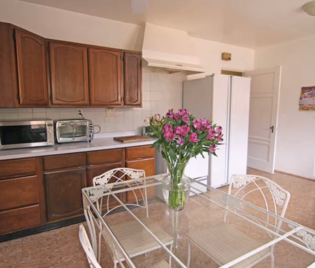 A kitchen with wooden cabinets, a white refrigerator, a microwave, a toaster oven, and a glass dining table with white metal chairs. A vase with purple flowers is placed on the dining table.