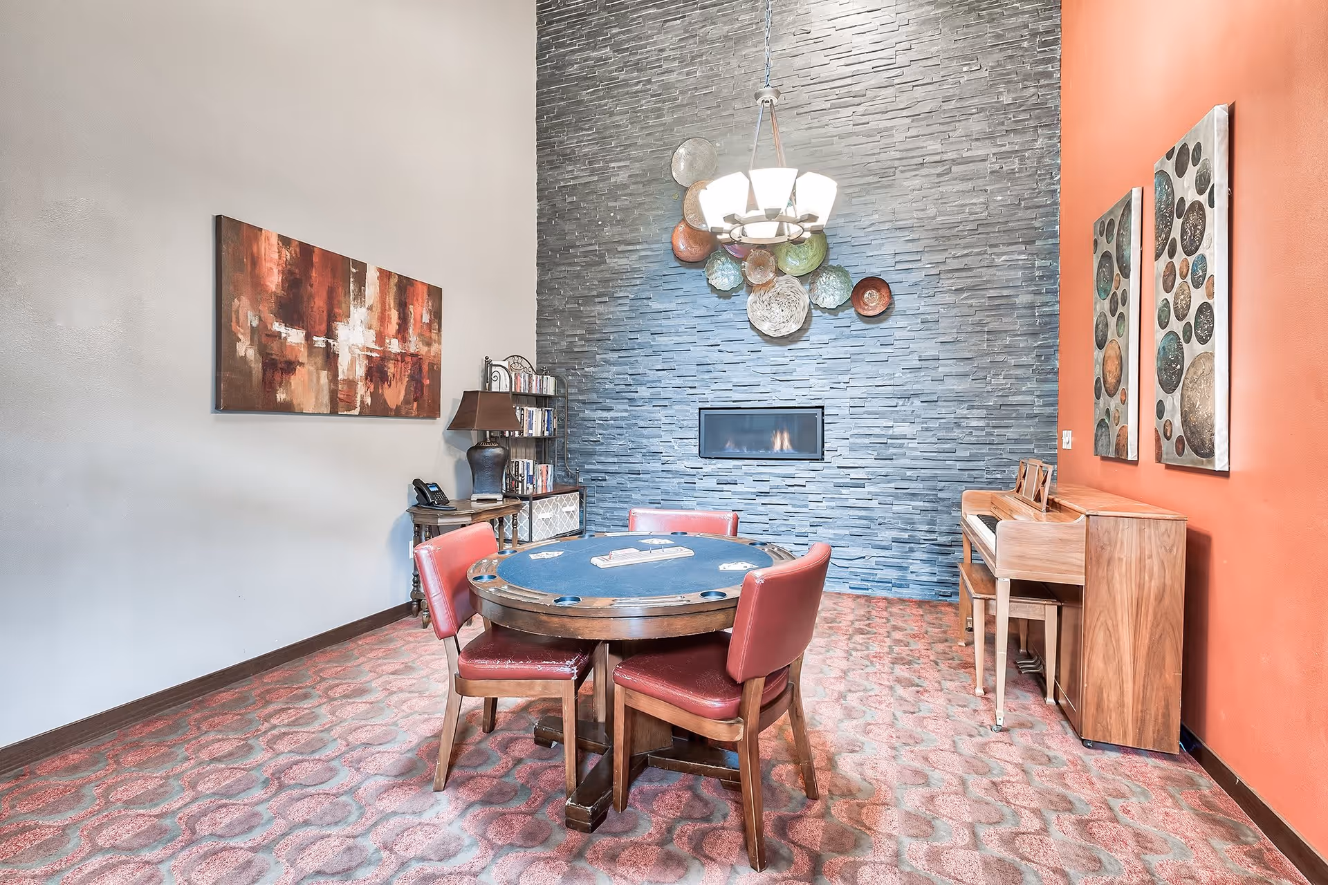 A cozy interior room with a round poker table surrounded by four red cushioned chairs. The room features a modern stone fireplace on a gray textured wall, colorful decorative plates above the fireplace, and two abstract paintings on an orange wall. There is a wooden piano with a matching bench on the right side and a small table with a lamp and books on the left side. The floor is carpeted with a patterned design.