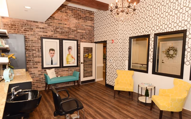 Interior view of a stylish room with wood flooring, a brick accent wall, and patterned wallpaper. The room features a teal bench with two framed portraits above it, two yellow patterned armchairs with a small round table between them, a black salon chair, a black sink, and a chandelier hanging from the ceiling.