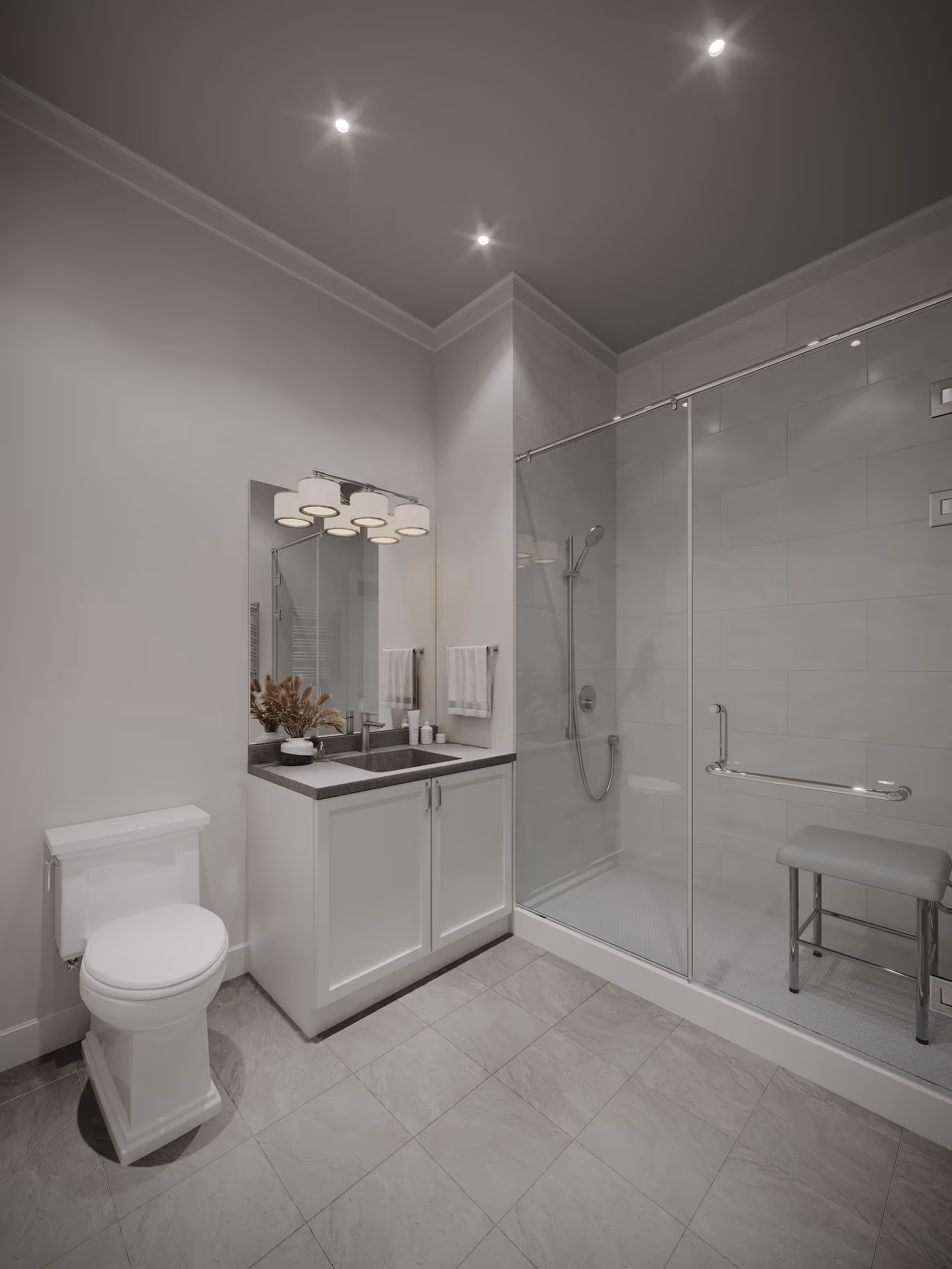 A modern bathroom featuring a white toilet, a vanity with a dark countertop and a large mirror above it, and a glass-enclosed shower with a handheld showerhead and a small bench inside. The floor is tiled in light gray, and the walls are painted white with recessed ceiling lights.