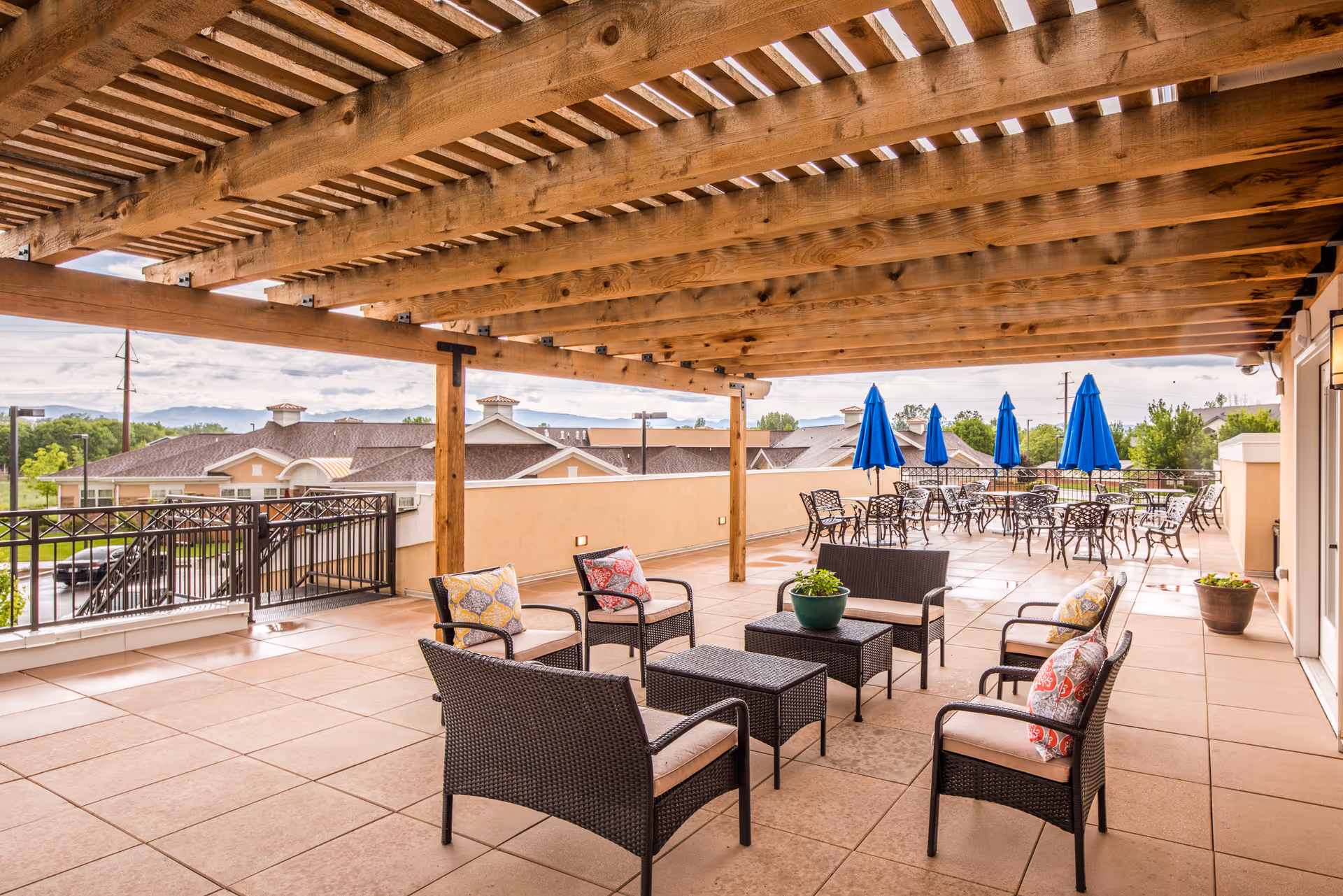 Outdoor patio area with a wooden pergola overhead, featuring wicker seating with cushions and small tables in the foreground. In the background, there are several metal tables with blue umbrellas. The patio overlooks nearby buildings and greenery under a partly cloudy sky.