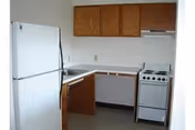 Small kitchen area with a white refrigerator on the left, wooden cabinets above and below a white countertop, a sink, and a white stove with an exhaust hood on the right.