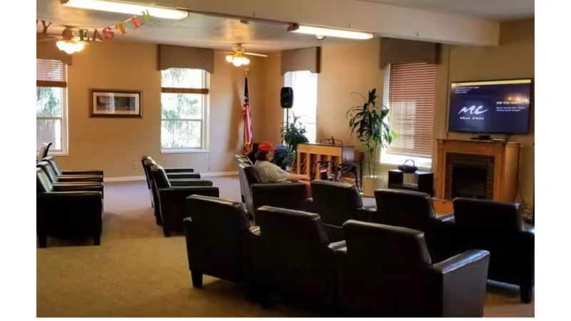 A common area in a senior living facility with several black armchairs arranged in rows facing a television mounted above a fireplace. A person wearing a red cap is seated in one of the chairs. The room has beige walls, windows with blinds, an American flag, potted plants, and ceiling fans with lights. A colorful 'EASTER' banner is hanging near the ceiling.