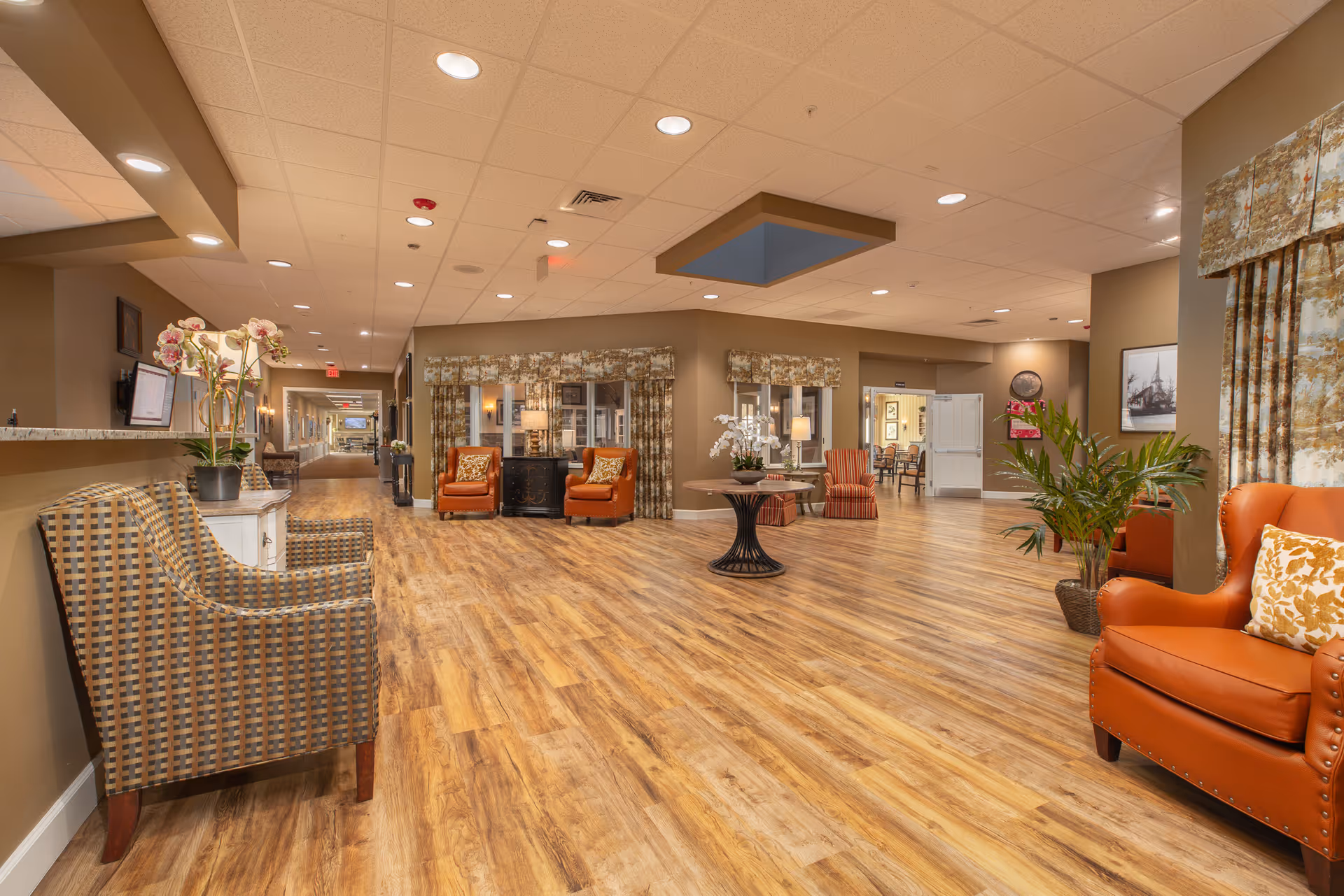 Spacious and well-lit interior common area of Cedarbrook Memory Care Community featuring wood flooring, comfortable armchairs in orange and patterned upholstery, a round table with a floral arrangement, potted plants, framed artwork on the walls, and large windows with patterned curtains.