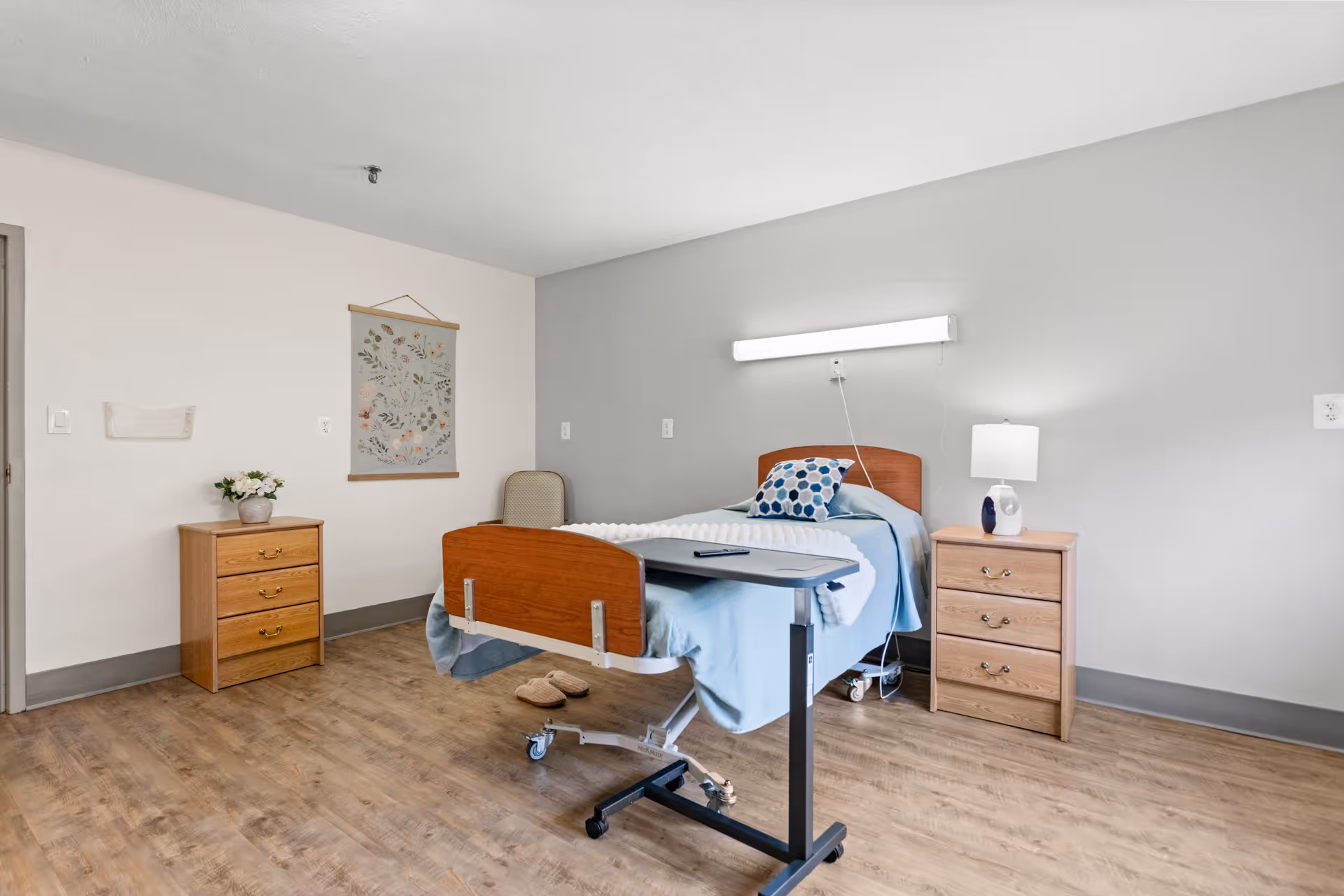 A simple nursing care bedroom with a single hospital bed covered with light blue bedding and a patterned pillow. There is a small overbed table with a remote on it, a wooden nightstand with a lamp, a wooden dresser with a small plant, a chair in the corner, and a floral wall hanging. The floor is wood and the walls are painted light gray and white.