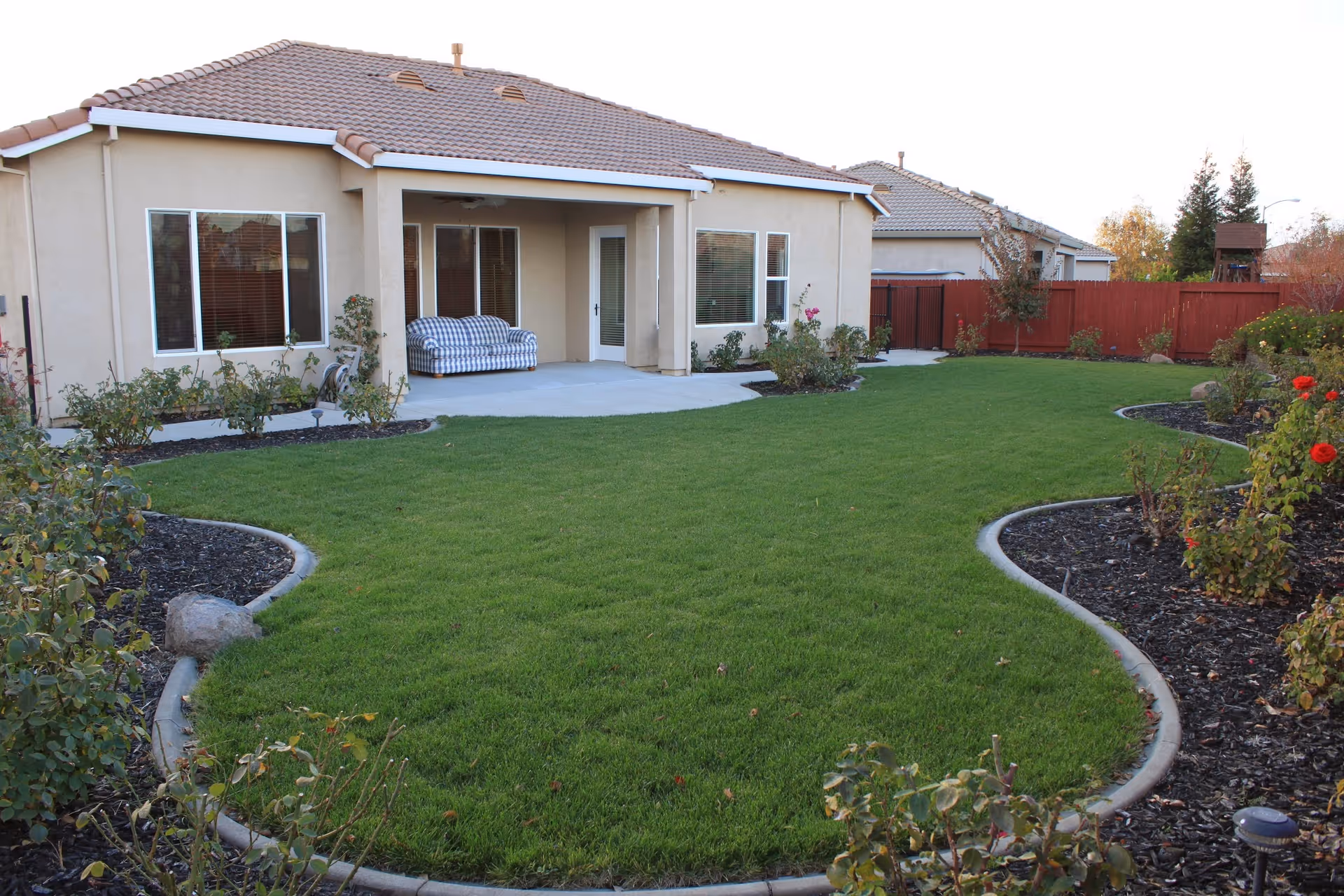 A well-maintained backyard with a green lawn, bordered by flower beds with various plants and flowers. A beige house with a tiled roof and large windows overlooks the yard. A covered patio area has a striped cushioned bench and a door leading inside.