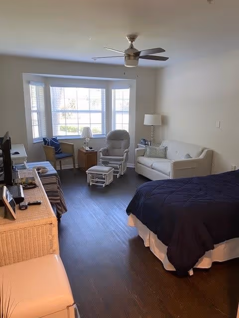 A bright and cozy bedroom with a large window letting in natural light. The room features a bed with a dark blue comforter, a white couch with pillows, a reclining chair with an ottoman, a small table with a lamp, and two chairs near the window. A ceiling fan is mounted above, and the floor is dark wood.