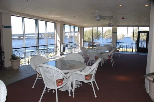 Indoor seating area with white wicker chairs and glass-top tables arranged on a carpeted floor. Large windows provide a view of a body of water and trees outside. Ceiling fans and recessed lighting are visible on the ceiling.