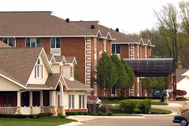 Brick multi-story senior living facility with a covered glass entry canopy, landscaped grounds, and nearby cottage-style units.