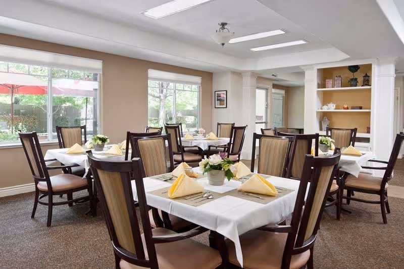 A bright dining room with several tables covered with white tablecloths, each set with yellow folded napkins, placemats, silverware, and small flower centerpieces. Large windows let in natural light and show greenery outside. The room has beige walls, carpeted floor, and wooden chairs with cushioned seats.