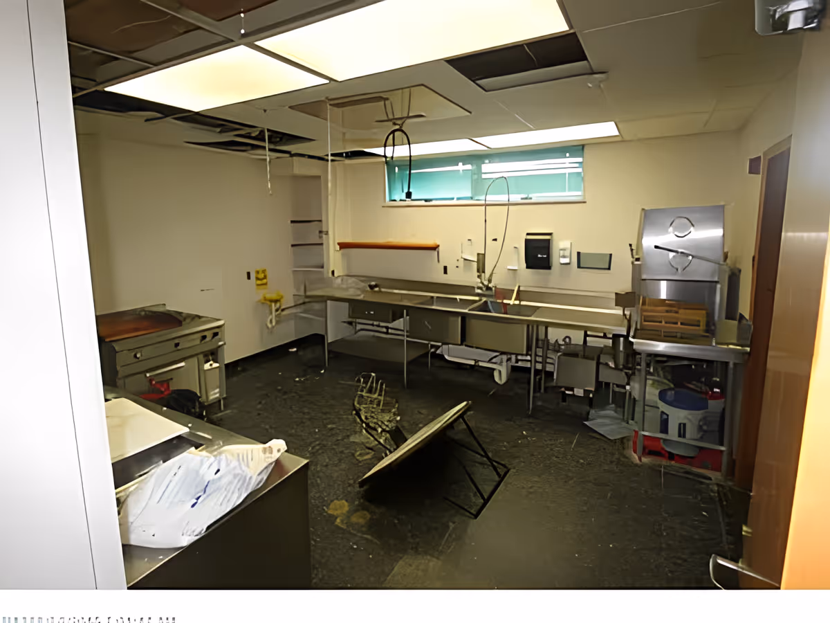 An abandoned and damaged commercial kitchen with broken ceiling tiles, a fallen table, stainless steel sinks and counters, and various kitchen equipment scattered around. The room has a small window with a green shade and a door on the right side.