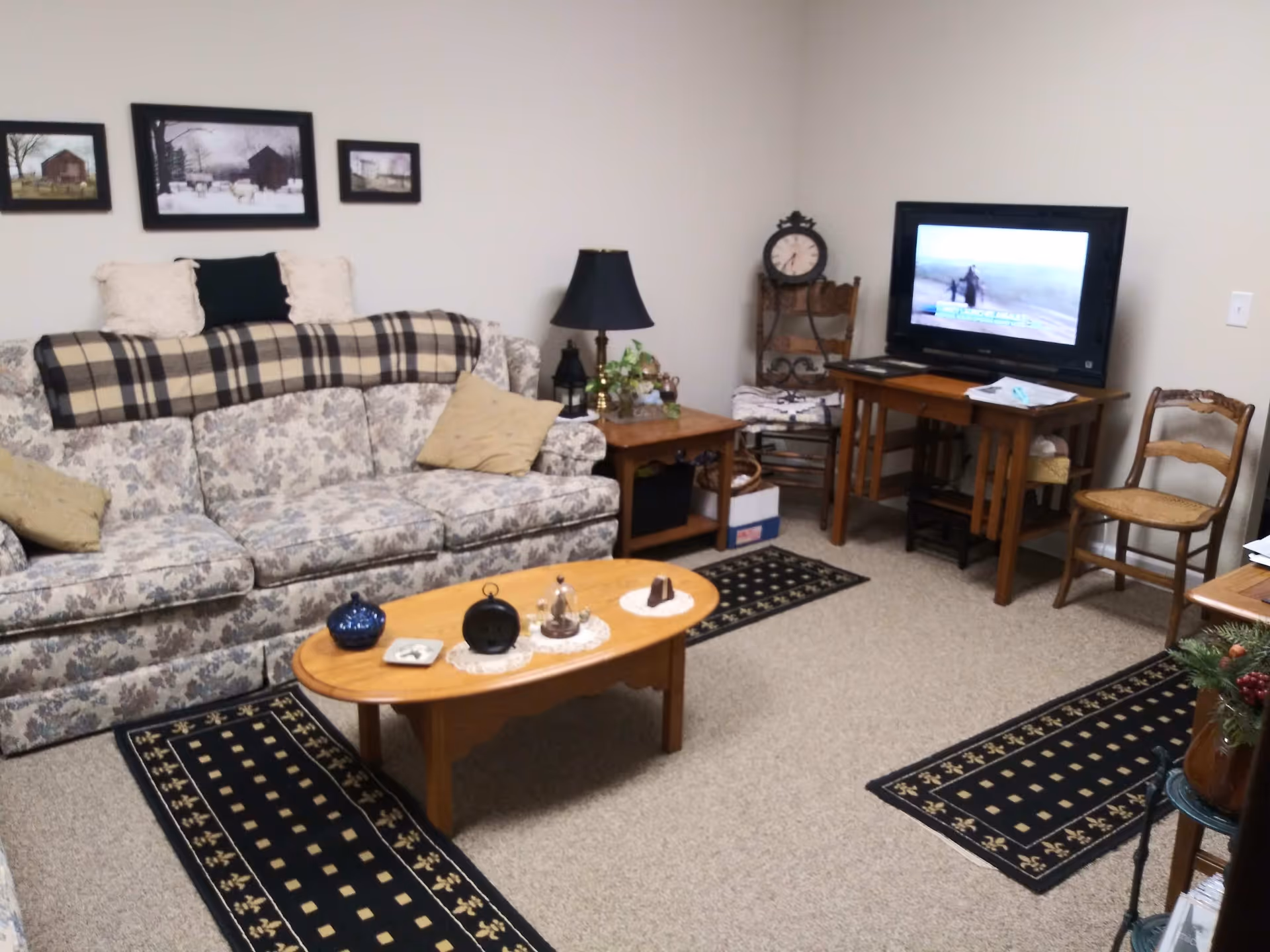 A cozy living room with a floral patterned sofa adorned with pillows and a plaid throw. A wooden coffee table with decorative items sits on a beige carpet with two black and gold patterned runners. There is a wooden side table with a lamp and plants, a TV on a wooden stand, and two wooden chairs. The walls are light-colored and decorated with three framed pictures.