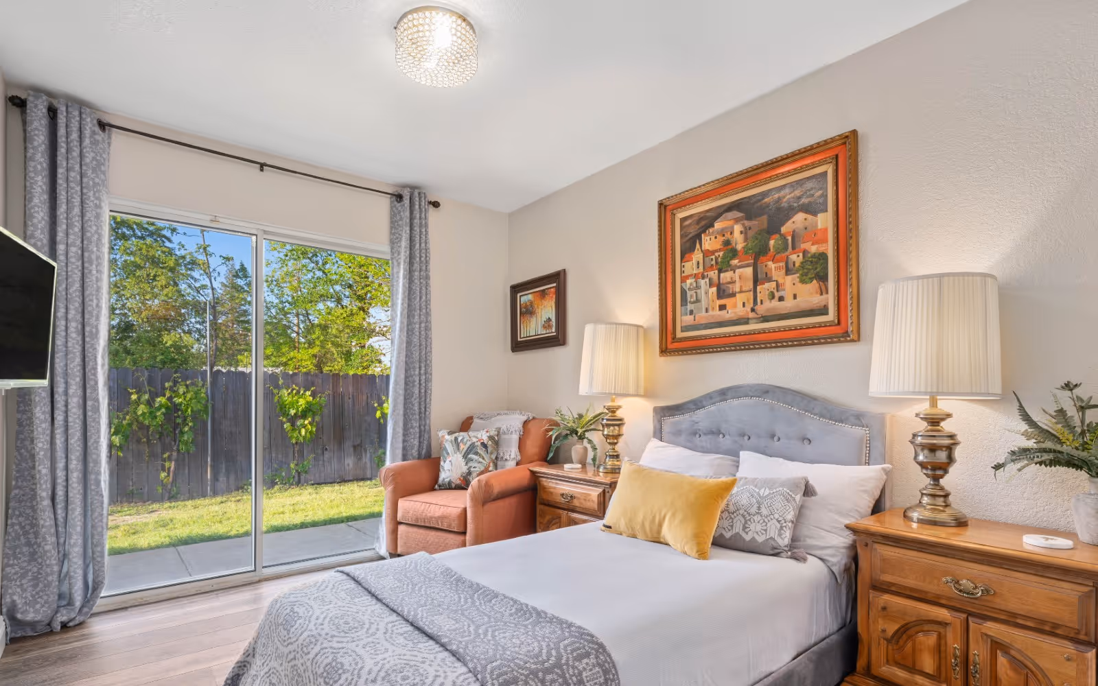 Bright bedroom with a bed, nightstands and lamps, an armchair by sliding glass doors opening to a backyard.