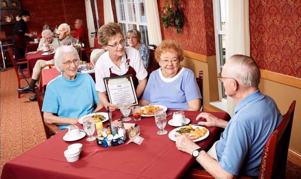 A group of elderly people sitting at tables with red tablecloths in a dining room. A staff member in a white shirt and maroon apron is showing a menu to two elderly women and a man seated at a table near a window with red patterned wallpaper. Other elderly people are seated at tables in the background.