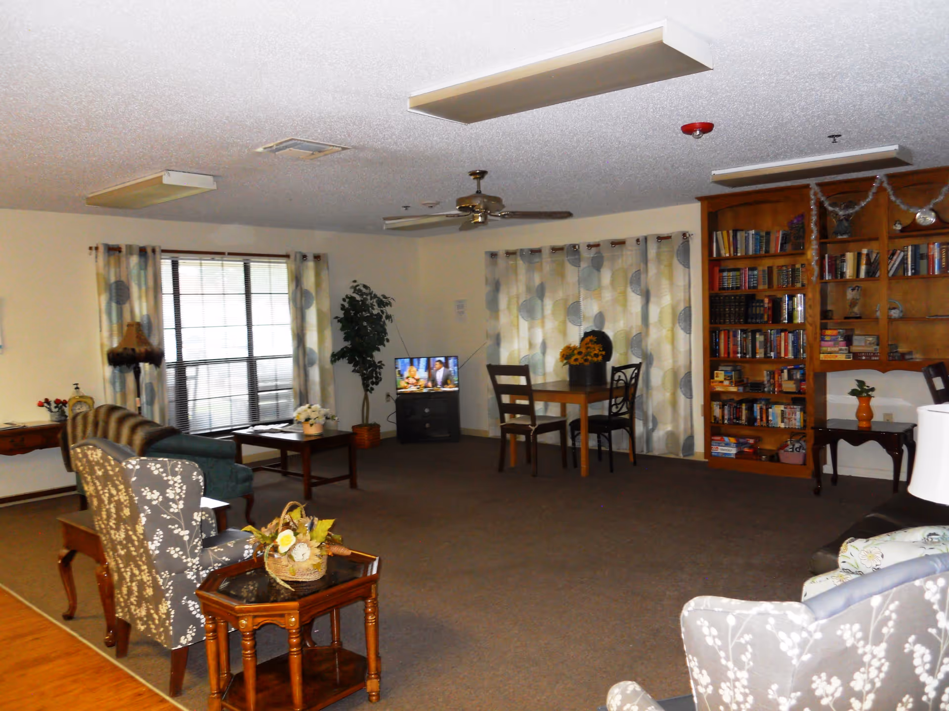A cozy living room area in an assisted living facility with patterned armchairs, a wooden coffee table, a small dining table with chairs, a TV on a stand, a tall bookshelf filled with books, and large windows with patterned curtains letting in natural light.