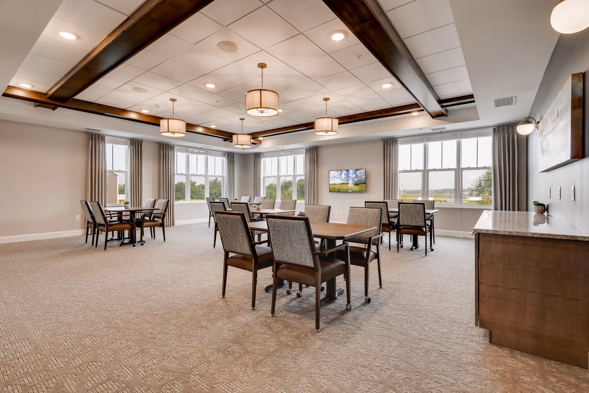 A spacious, well-lit dining room with multiple wooden tables and chairs arranged neatly. Large windows with curtains allow natural light to fill the room, and a flat-screen TV is mounted on the far wall. The ceiling features recessed lighting and decorative wooden beams with hanging light fixtures. There is a countertop with cabinets on the right side of the room.