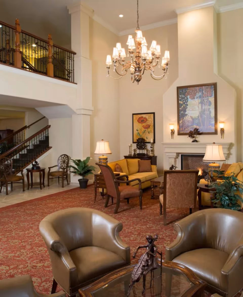 A spacious and elegant living room area with high ceilings, a large chandelier, and a fireplace. The room features comfortable seating including leather armchairs and upholstered chairs around a glass coffee table with a decorative sculpture. There are yellow sofas, side tables with lamps, framed artwork on the walls, and a red patterned carpet covering the floor. A staircase with wooden railings is visible in the background.