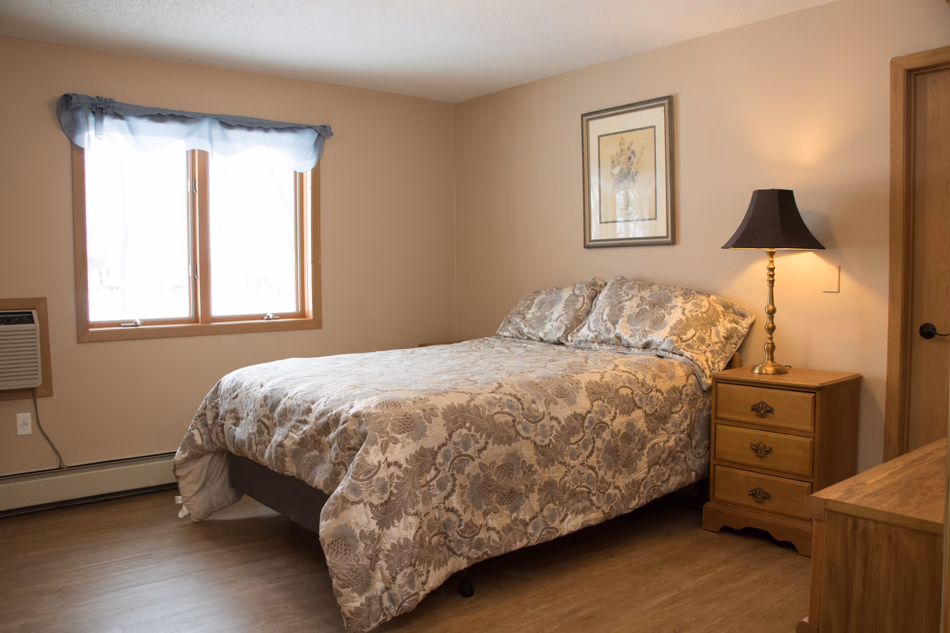 A bedroom with a double bed covered in a floral patterned comforter and matching pillows. There is a wooden nightstand with a lamp beside the bed, a framed picture hanging on the wall above the bed, a window with a blue valance, and a wooden door to the right.