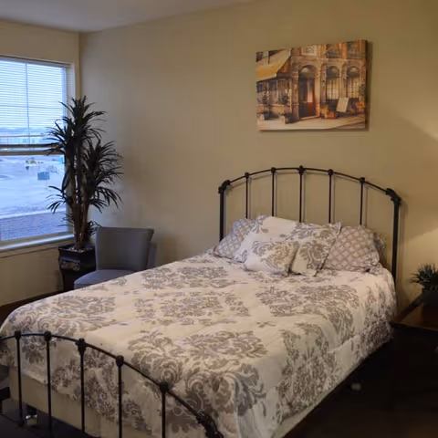 A bedroom with a metal framed bed covered with a white and gray patterned comforter and several pillows. There is a small gray chair and a tall potted plant near a window with blinds. A framed picture hangs on the beige wall above the bed.