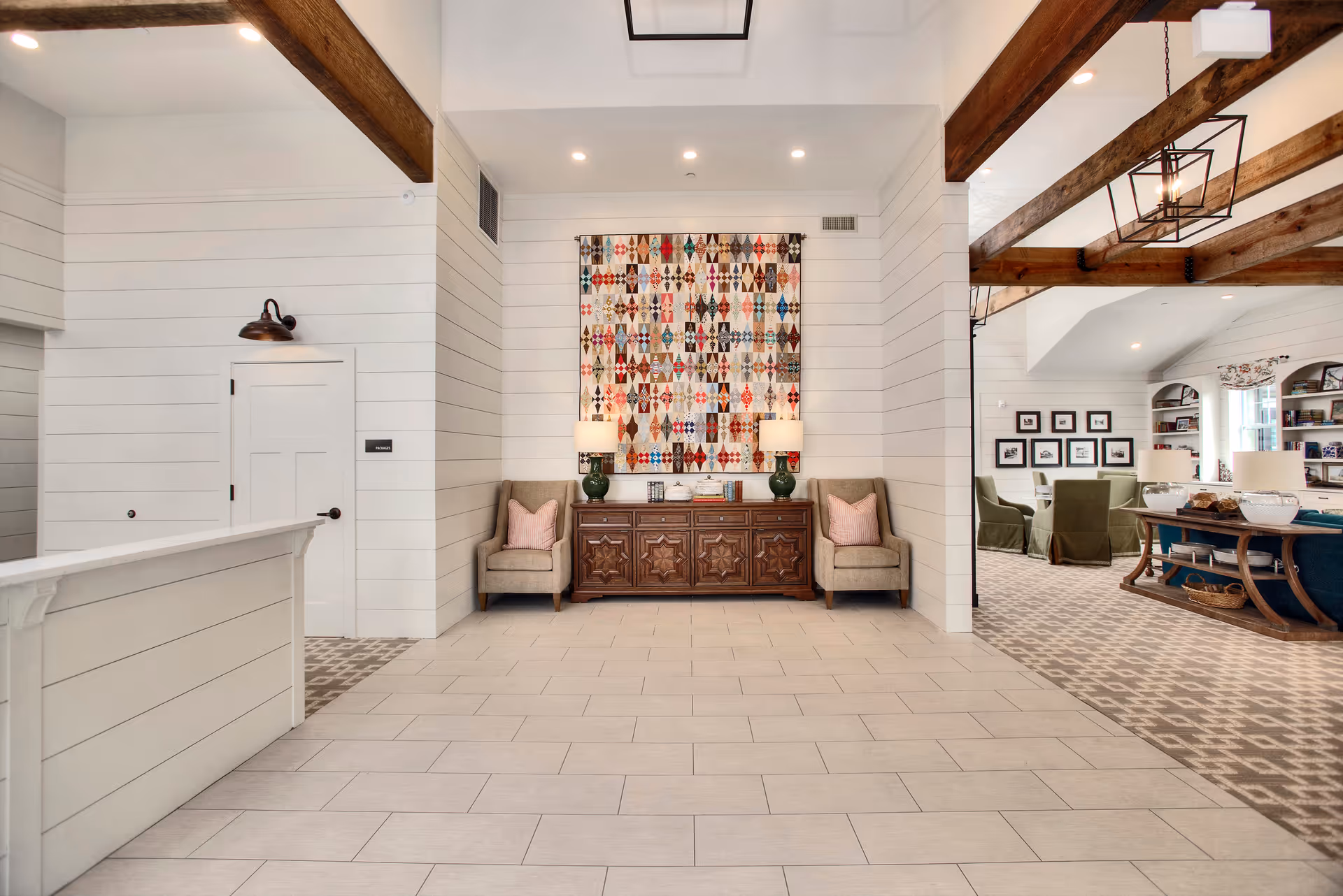 A bright and spacious interior area with white shiplap walls and exposed wooden ceiling beams. The center features a wooden sideboard with two green lamps and a colorful quilt hanging above it. Two beige armchairs with pink pillows flank the sideboard. To the right, there is a carpeted seating area with green chairs, framed pictures on the wall, and built-in bookshelves. The floor in the foreground is tiled in a light color.