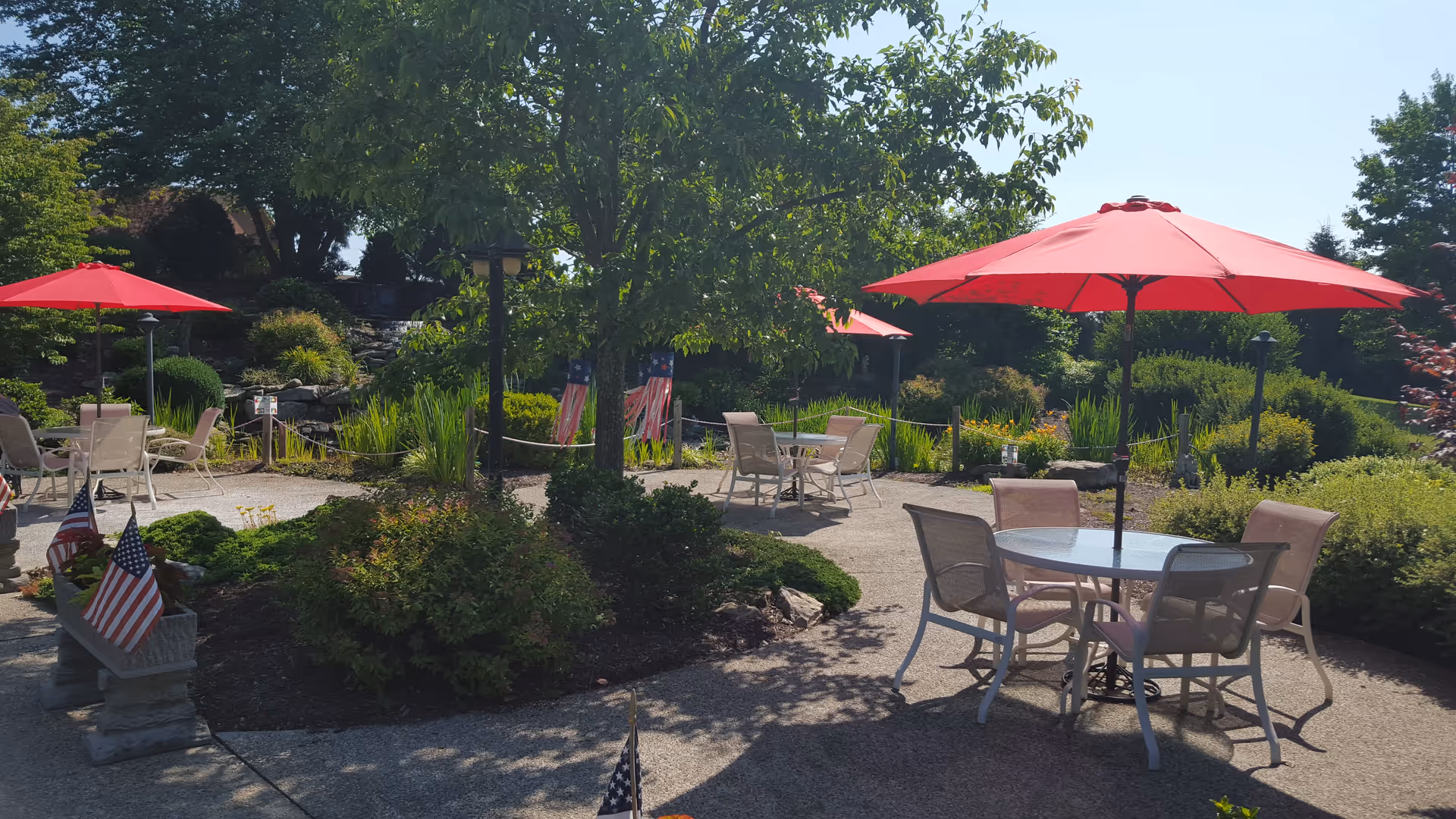 Outdoor garden area with several round tables and chairs under red umbrellas. The space is surrounded by lush greenery, bushes, and trees, with American flags placed in planters and along the pathway. The sky is clear and sunny.