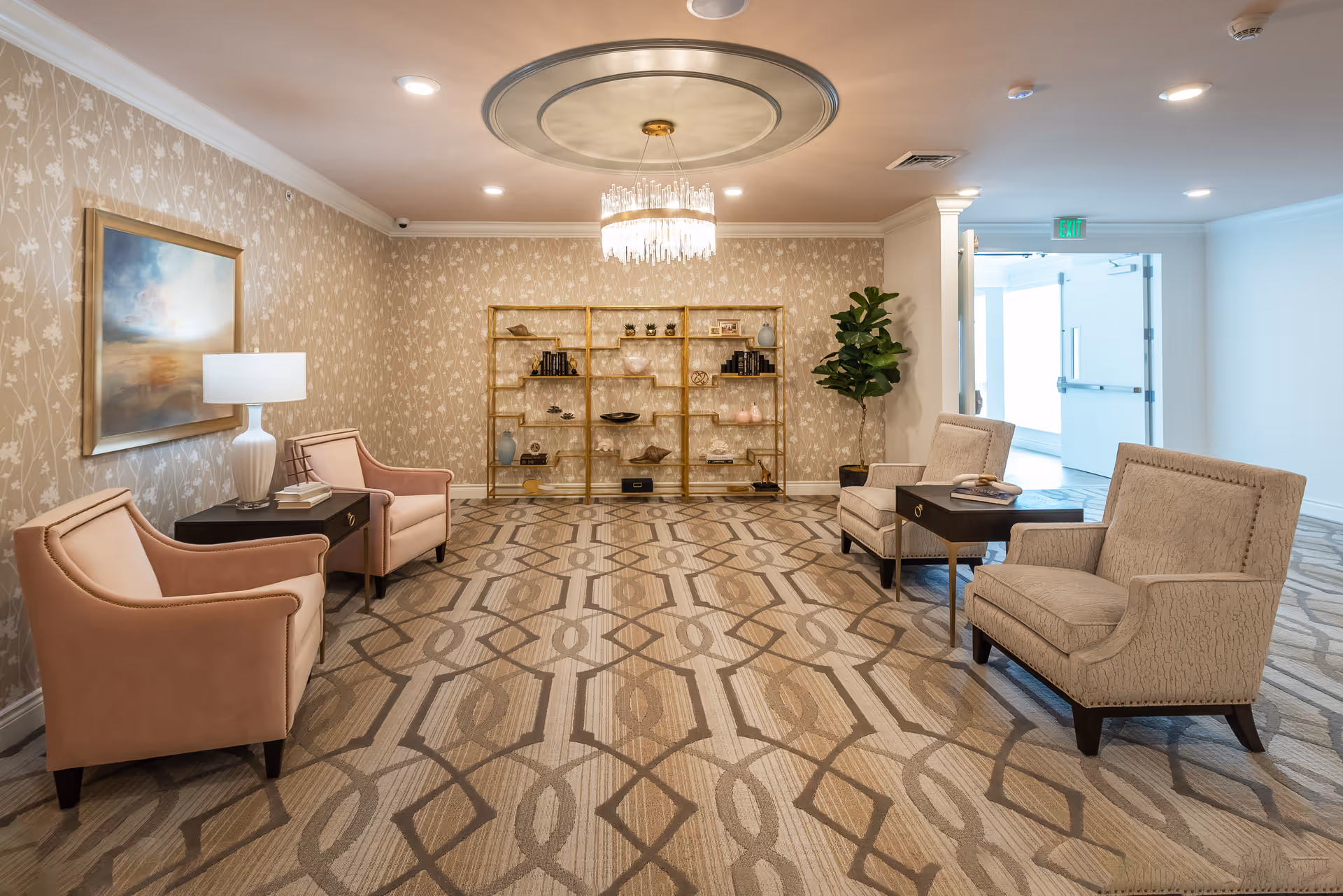 A spacious furnished lounge with pink and beige armchairs, side tables, a gold shelving unit, and a chandelier.