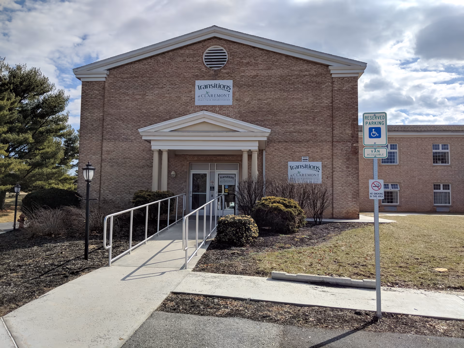 Front exterior view of a brick building with a ramp leading to the entrance. The building has signs that read 'Transitions at Claremont' and there is a reserved parking sign for handicapped access near the sidewalk. The sky is partly cloudy and there are some bushes and trees around the building.