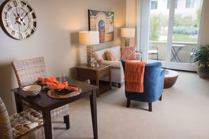 A cozy living room area with a small dining table set for two, featuring wicker chairs with cushions and orange napkins. The room includes a beige sofa with patterned pillows, a blue armchair with an orange throw, two table lamps on wooden side tables, a large wall clock, and a framed picture on the wall. A glass door leads to an outdoor patio with a small table and chairs, and there is a potted plant near the door.