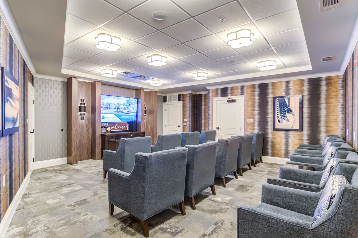 A cozy media room with two rows of gray upholstered armchairs facing a large flat-screen TV mounted on a wooden accent wall. The room features patterned wallpaper, modern ceiling lights, and framed artwork on the walls.
