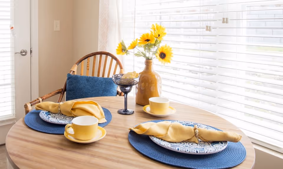 A small round wooden dining table set for two with blue placemats, patterned plates, yellow napkins with napkin rings, and yellow cups and saucers. A vase with bright yellow sunflowers and a small blue glass bowl with snacks are on the table. The table is near a window with white blinds, and a wooden chair with a blue cushion is visible.