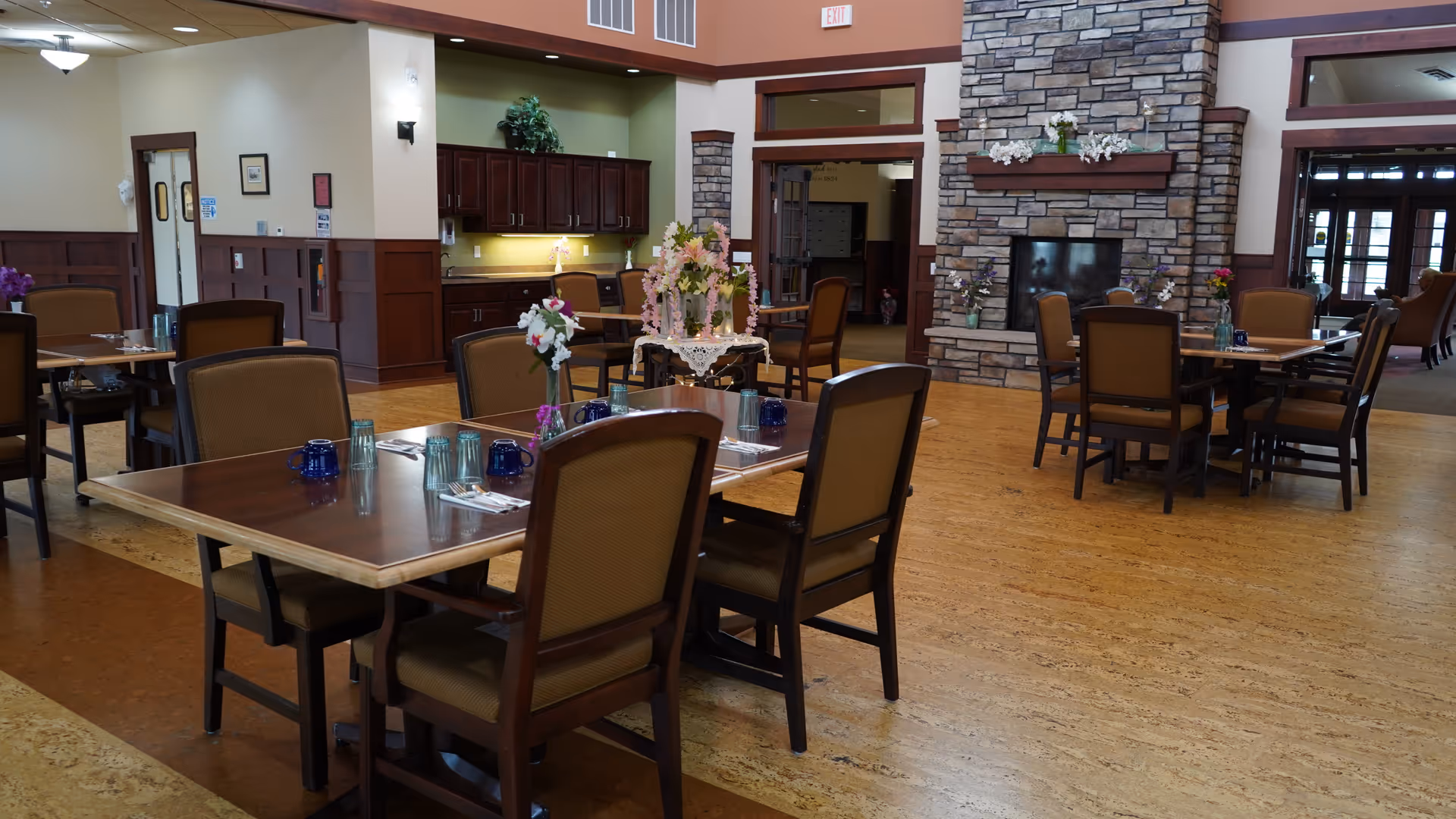A spacious dining area in a senior living facility with several wooden tables and cushioned chairs arranged neatly. Each table is set with blue and clear drinking glasses, silverware, and small floral centerpieces. The room features a stone fireplace with decorative flowers on the mantel, wood paneling on the walls, and a warm, inviting atmosphere. In the background, there is a kitchenette area with cabinets and a countertop, and large windows letting in natural light.
