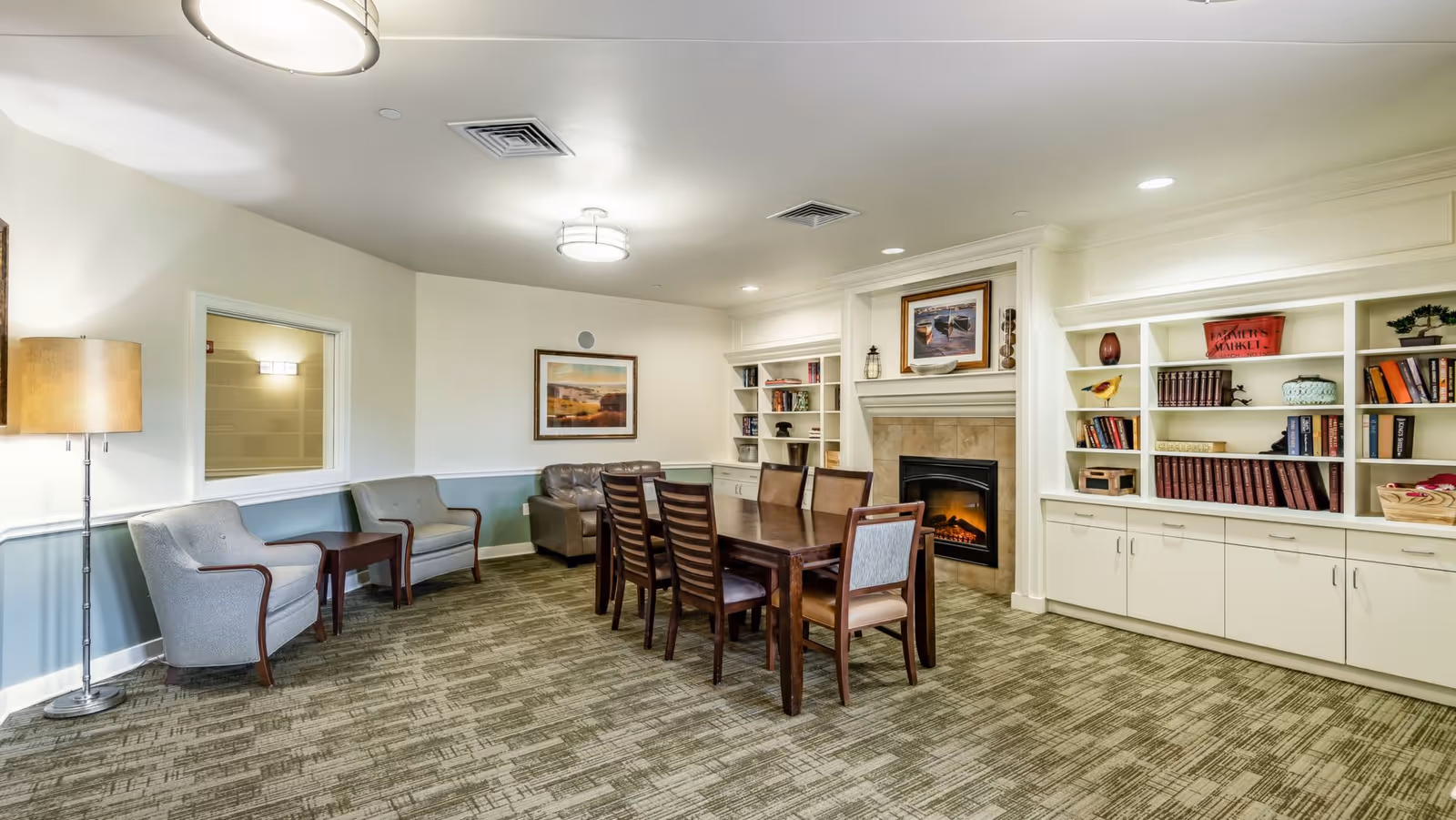 A well-lit common area with a long wooden table surrounded by eight chairs, two gray armchairs with a small table between them, a fireplace with a framed picture above it, and built-in white bookshelves filled with books and decorative items.