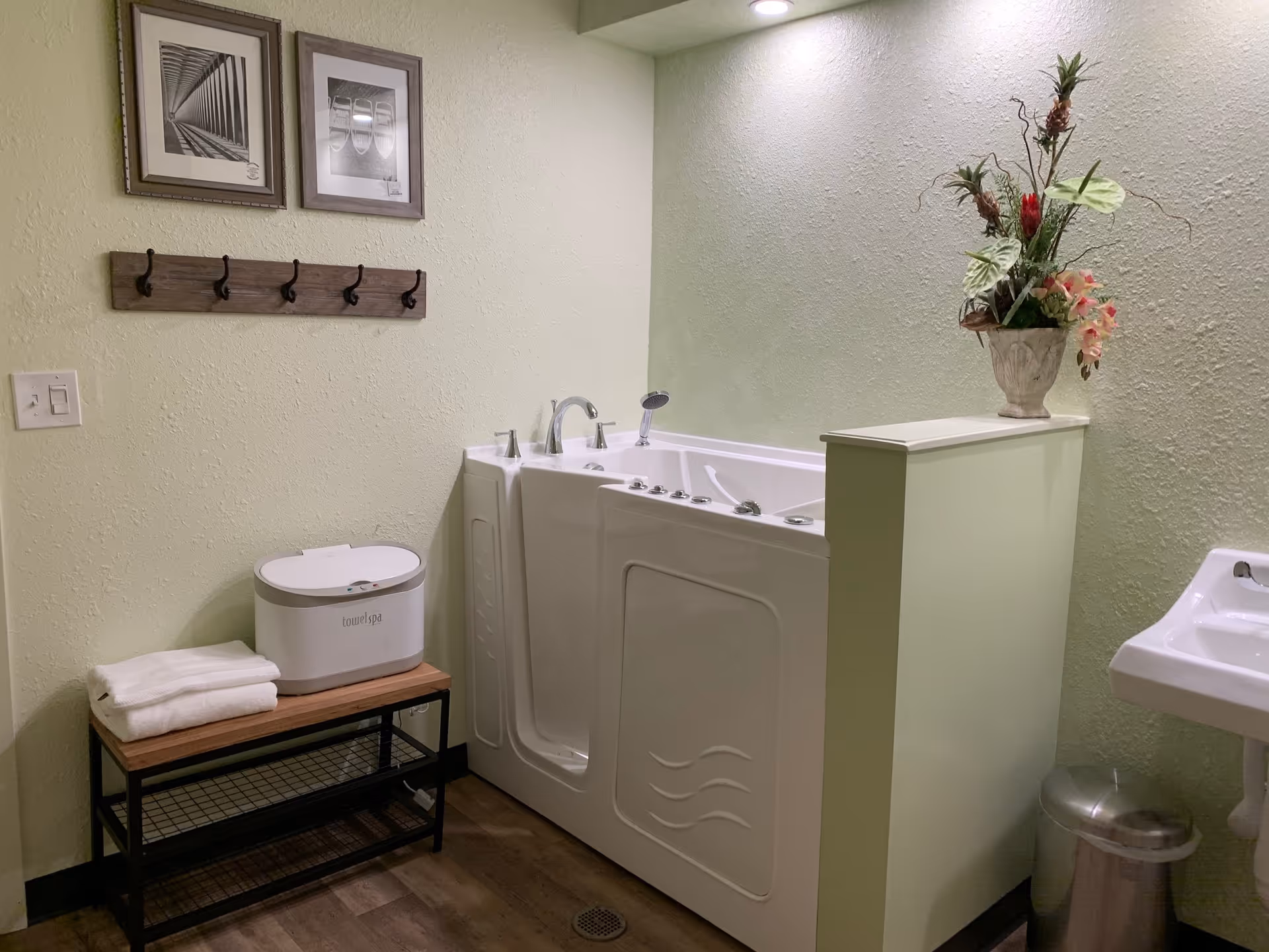 A bathroom with a walk-in bathtub featuring multiple jets and a handheld showerhead. To the left, there is a wooden bench with folded white towels and a towel spa device on top. Above the bench, two framed black and white pictures and a wooden coat rack with hooks are mounted on a light green textured wall. To the right, a white pedestal sink and a silver trash can are visible. A decorative flower arrangement is placed on a half wall next to the bathtub.