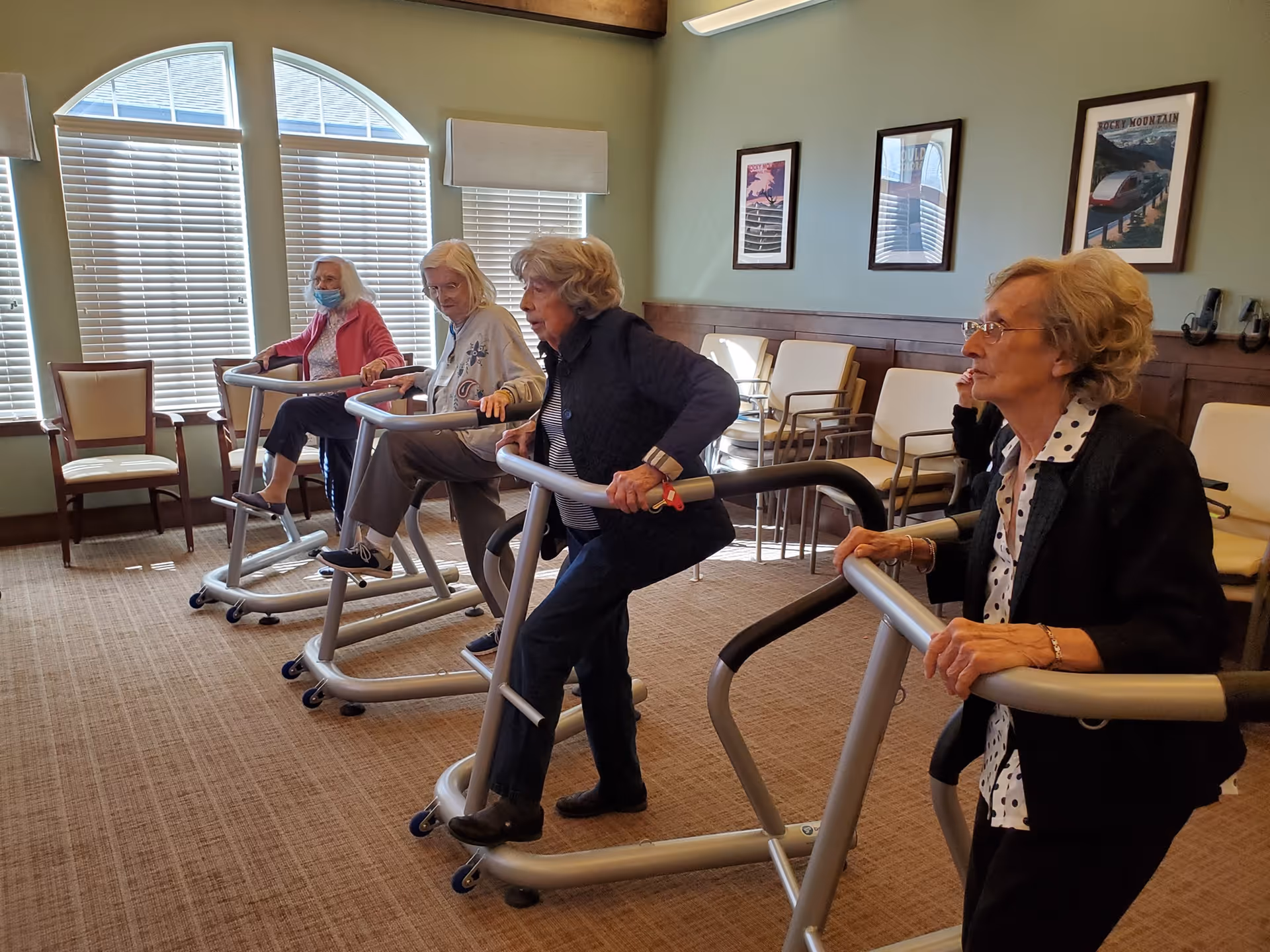 Four elderly women using standing exercise equipment in a well-lit room with large windows and several chairs arranged along the walls. The women appear focused on their activity, and one is wearing a face mask.