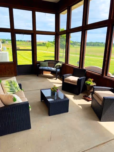 A bright sunroom with large windows offering a view of a green lawn and trees outside. The room is furnished with dark wicker chairs and a loveseat with beige cushions and green leaf-patterned pillows. A glass-top coffee table with a small plant is in the center, and there is a wooden bench with cushions near the back door.
