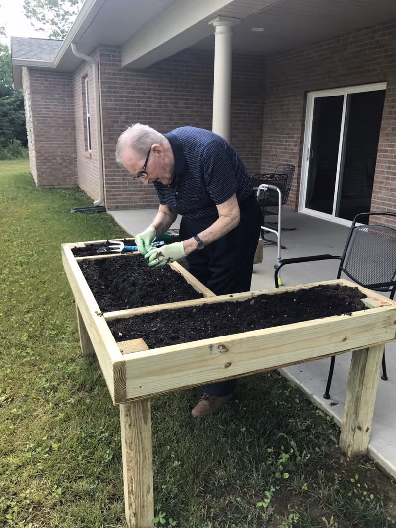 An elderly man wearing gloves is gardening at a raised wooden planter filled with soil outside a brick building with a covered patio. There are chairs and a walker on the patio.