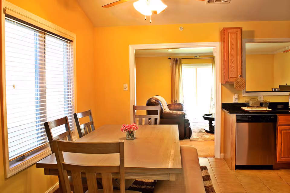 Bright dining area with a wooden table, chairs, and open kitchen in warm yellow.
