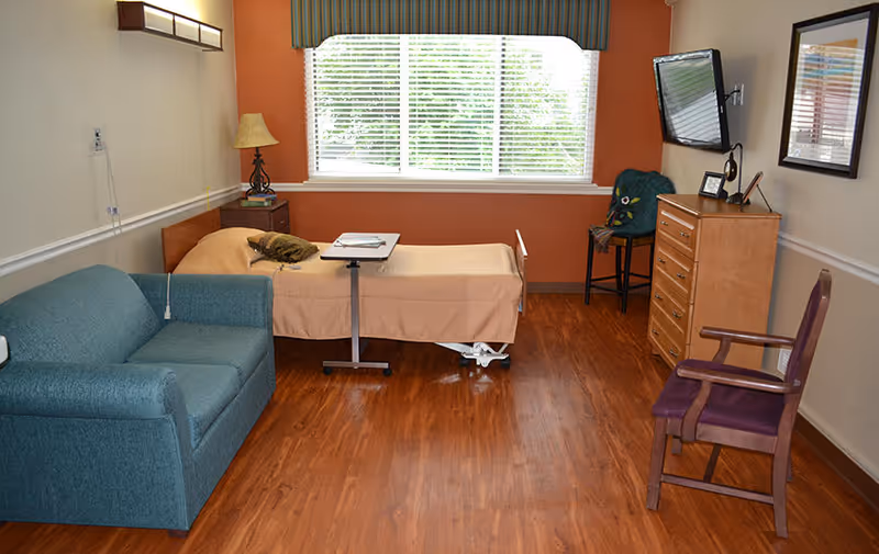 A nursing facility room with a single bed covered in beige bedding, a green sofa, a wooden dresser with a flat-screen TV mounted above it, a wooden chair with purple cushion, and a window with blinds and a valance. The floor is wooden and the walls are painted beige and orange.