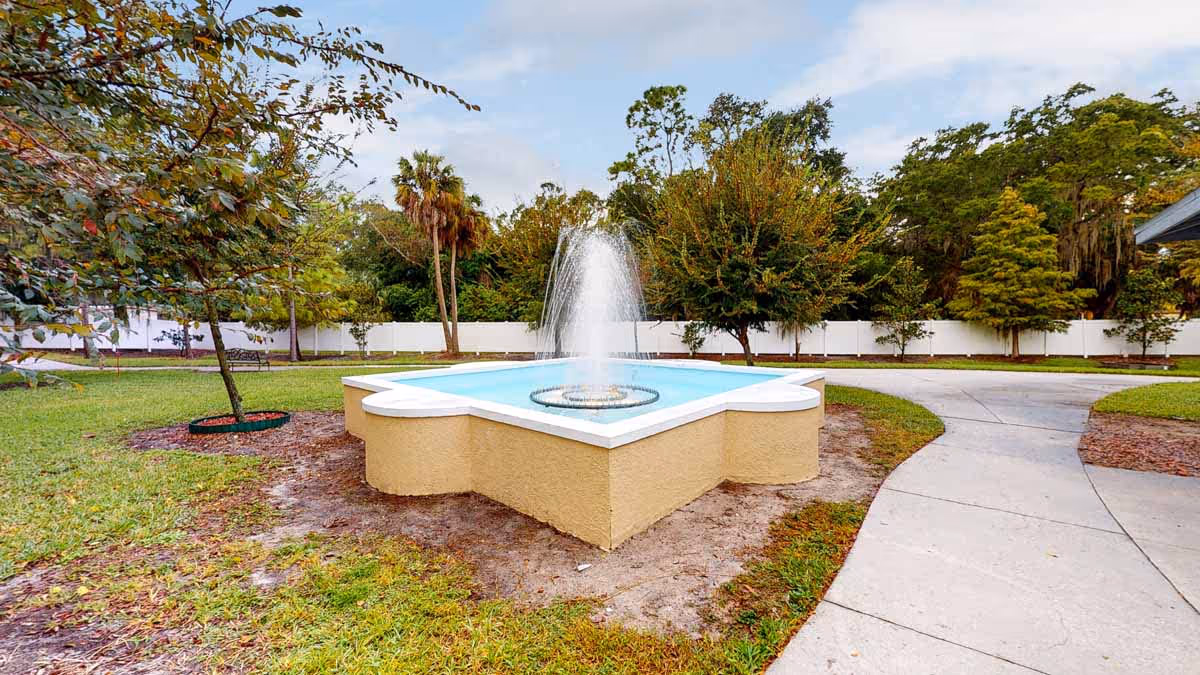 Outdoor courtyard with a central raised decorative fountain, surrounding grass, trees, and paved walkways.