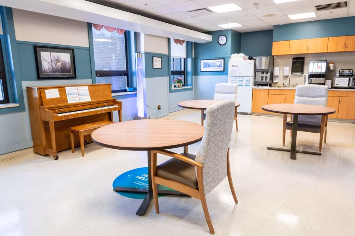 Bright communal dining/activity room with round tables and chairs, a piano against the wall, and a kitchenette area.