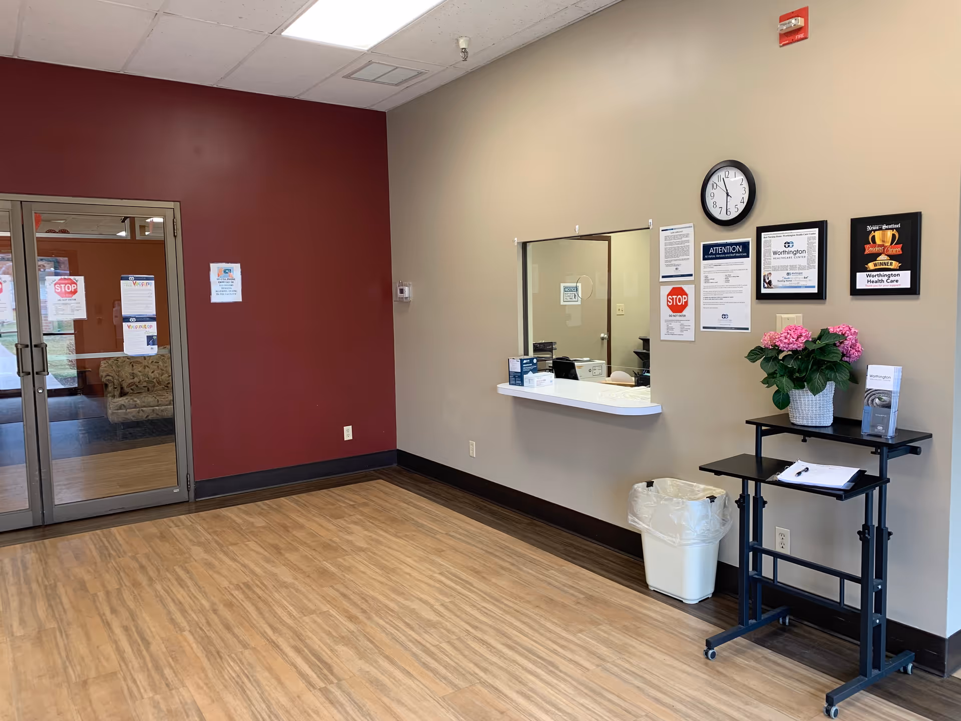 Reception area of Worthington Healthcare Center with a service window, a clock on the wall, informational posters, a small table with a pink flower arrangement and brochures, a trash can, and double glass doors leading to another room with a couch visible.
