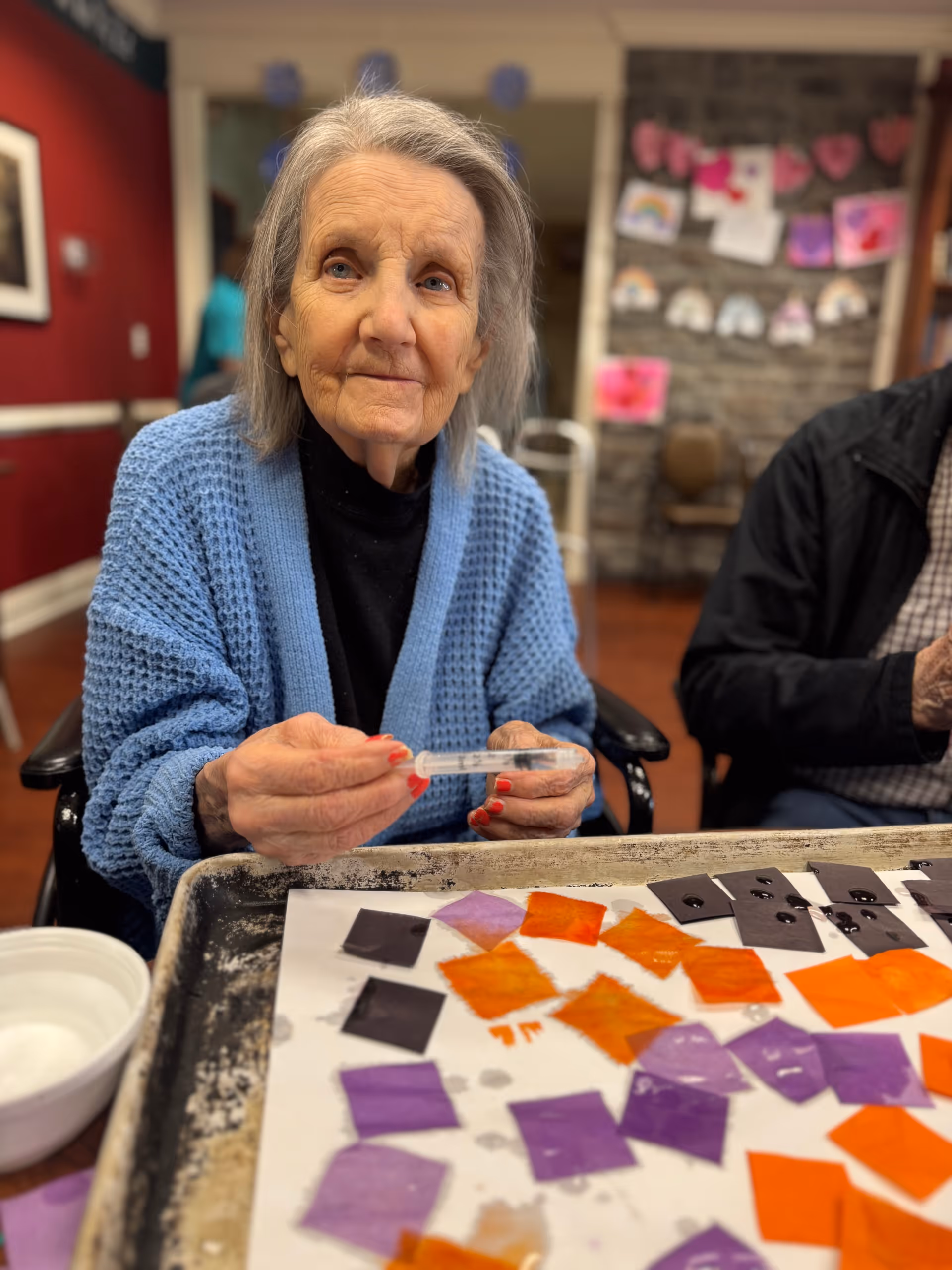 An elderly woman wearing a blue knitted cardigan and black shirt is seated at a table engaged in an arts and crafts activity. She is holding a small plastic dropper above a tray with various colored paper squares in orange, purple, and black. The background shows a room with red walls, a stone wall with children's artwork, and another person partially visible sitting beside her.