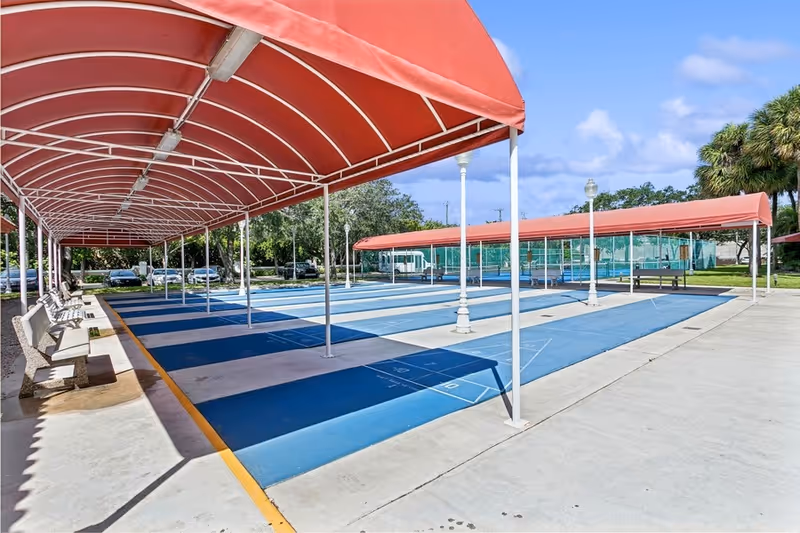 Outdoor shuffleboard courts with blue playing surfaces and red canopies overhead. There are benches along the left side and trees and parked cars in the background under a partly cloudy sky.