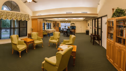 A spacious common area in a senior living facility with green carpet and beige walls. The room features several green upholstered armchairs arranged around wooden side tables, a wooden cabinet with glass doors containing books, and large windows with floral valances letting in natural light. In the background, there is a dining area with tables and chairs, and some decorative plants on top of cabinets.