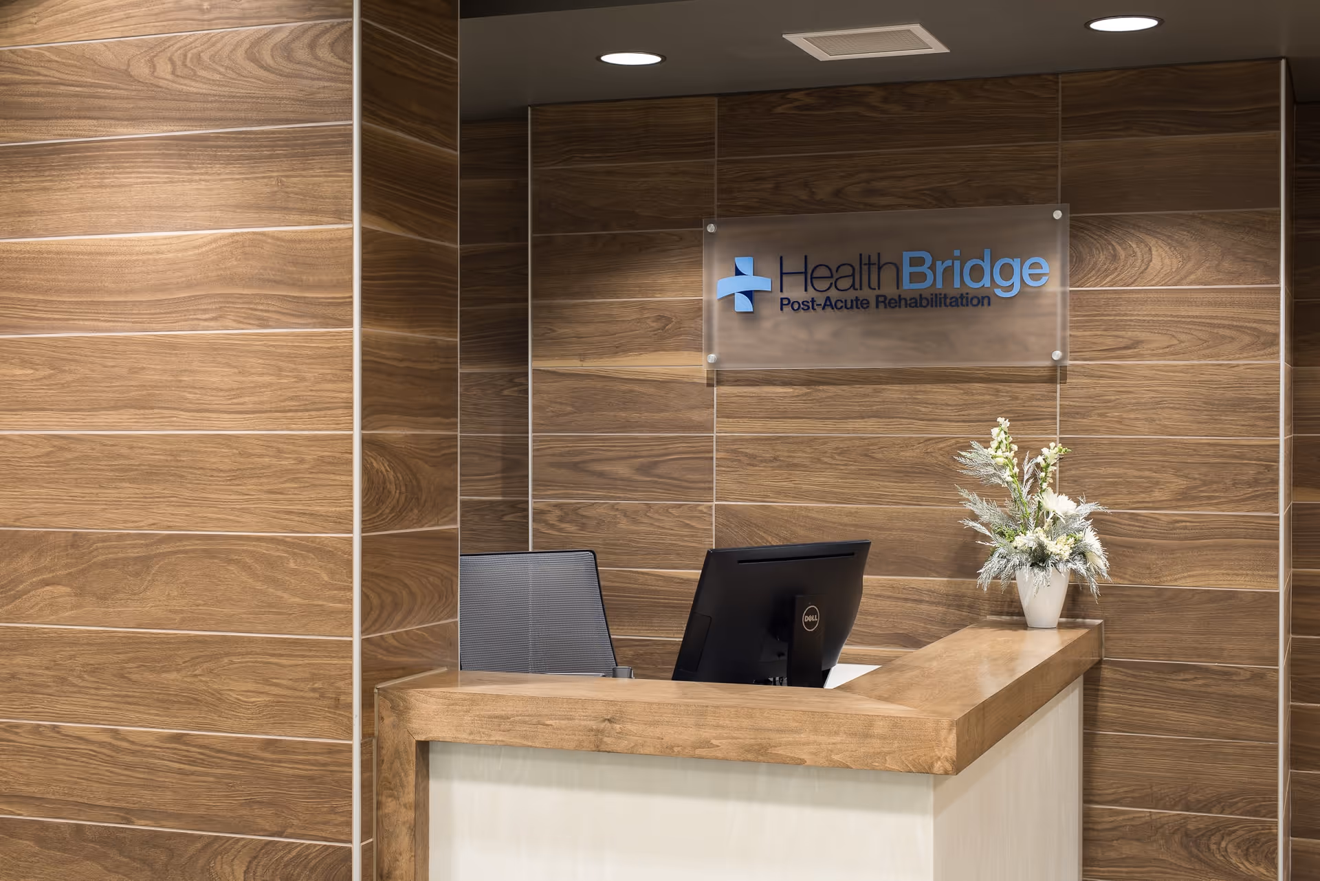 Reception desk area with wood-paneled walls, a computer monitor, a chair, and a vase with flowers. A sign on the wall reads 'HealthBridge Post-Acute Rehabilitation.'