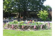 Circular raised stone flowerbed decorated with small American flags in a grassy courtyard with trees and a building in the background.