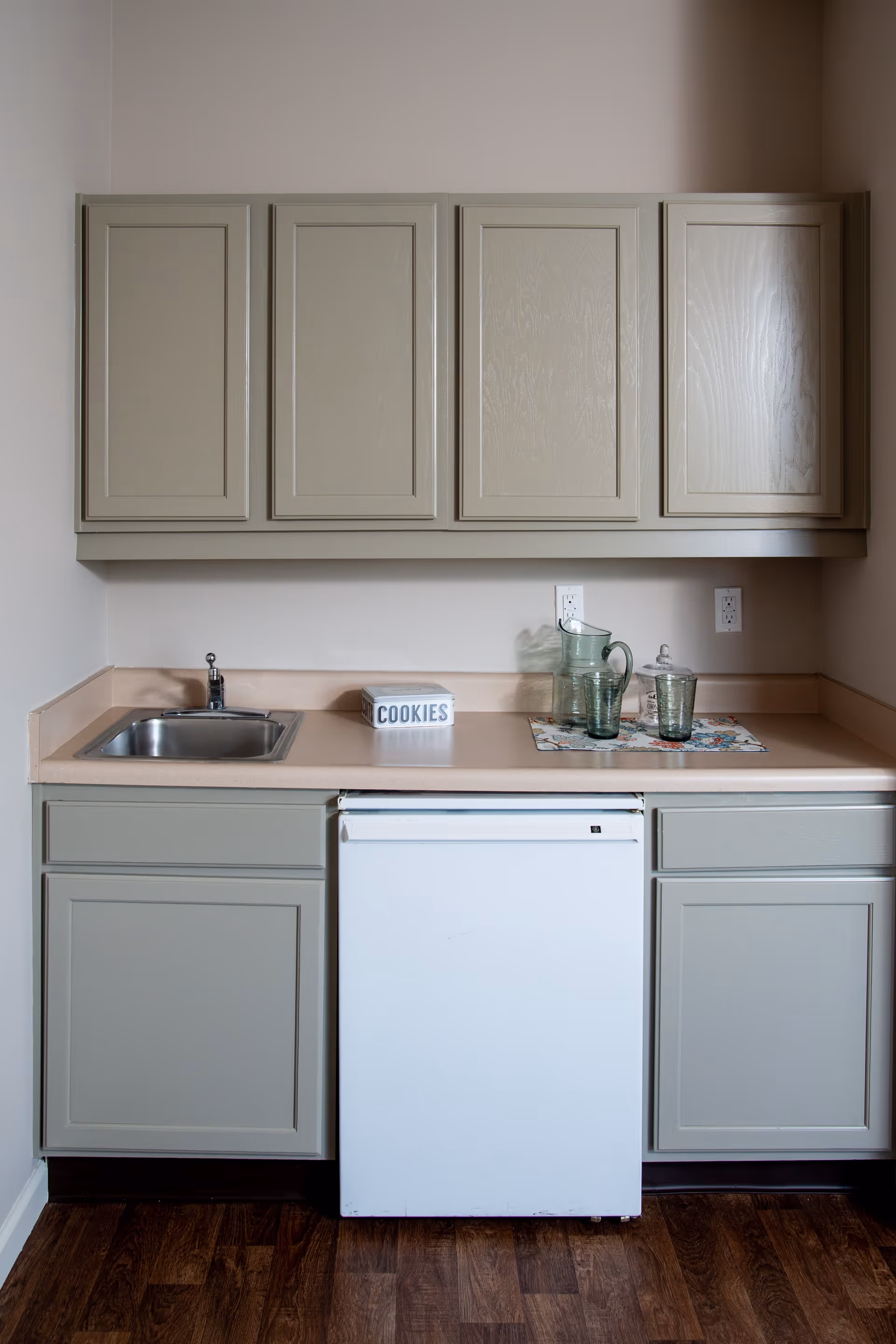 A small kitchen area with beige cabinets above and below a beige countertop. There is a small stainless steel sink on the left, a white mini refrigerator in the center, and a cookie jar labeled 'COOKIES' on the counter. To the right, there is a glass pitcher and two glasses on a floral placemat. The floor is dark wood.