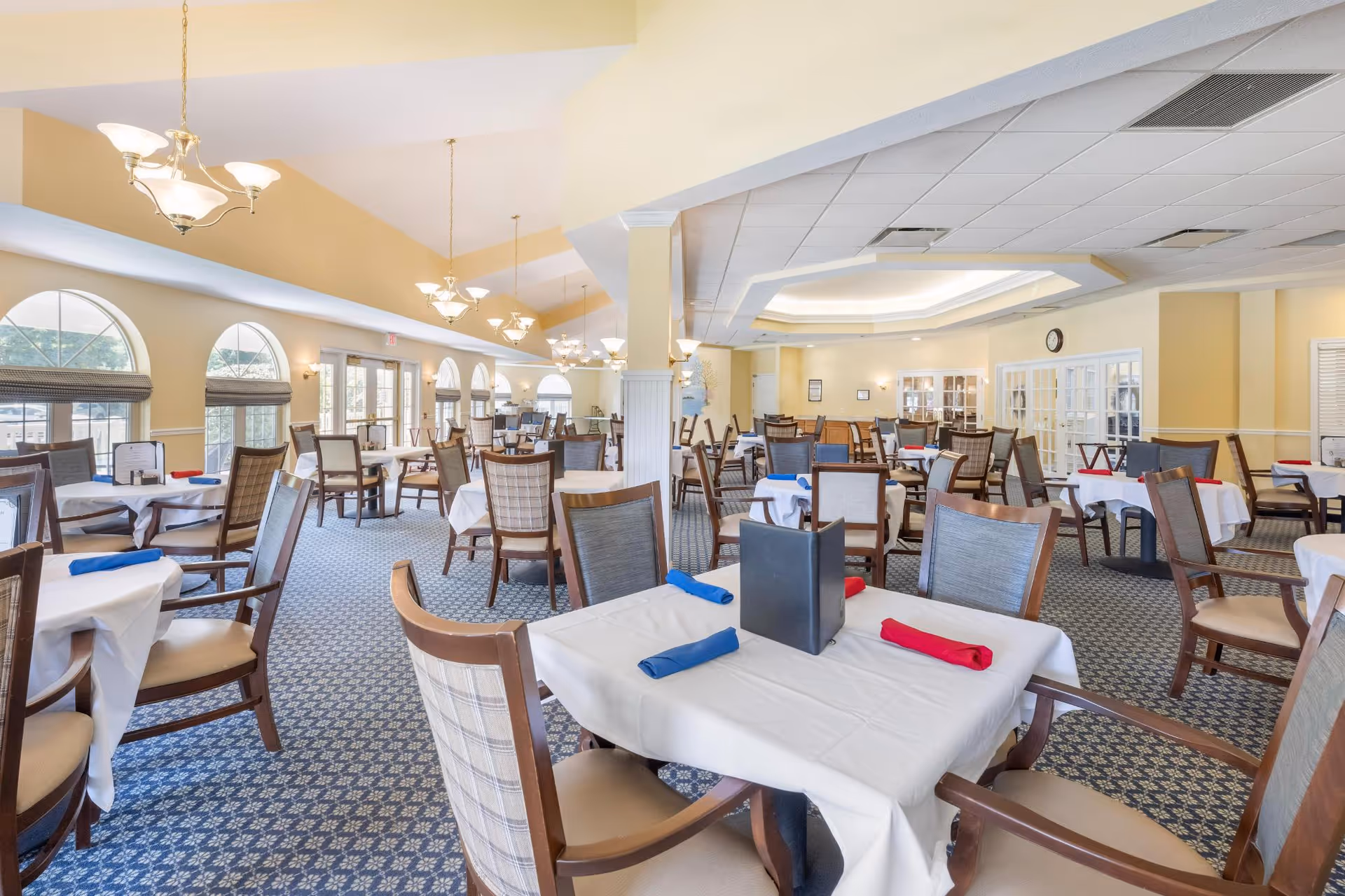 A spacious dining room with multiple tables covered in white tablecloths, each set with red and blue napkins. The room features large arched windows allowing natural light to fill the space, patterned carpet flooring, and elegant chandeliers hanging from the ceiling. Wooden chairs with cushioned seats surround each table.